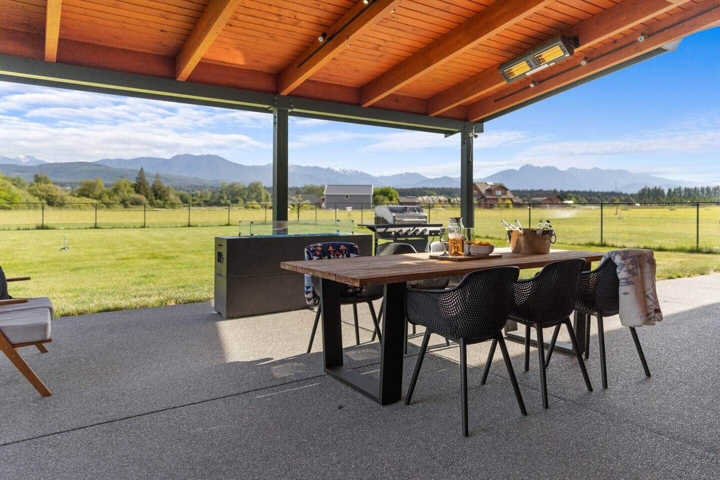 Een terras buiten met een houten tafel en stoelen, een barbecue en uitzicht op bergen.