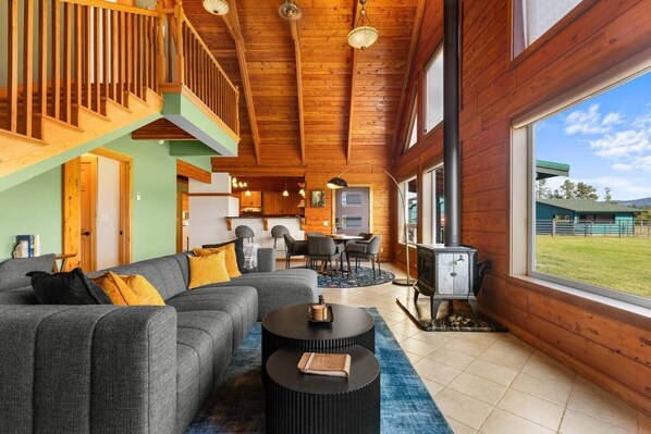 A spacious living area with a sofa, coffee table, and a fireplace. A staircase leads to an upper level, and there's a view of the outdoors through large windows.