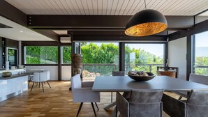 Una cuina moderna amb una illa gran, una zona de dinar amb taula i cadires, i grans finestres que ofereixen vistes a la vegetació.