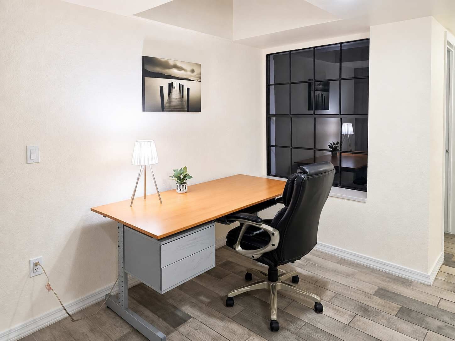 A modern office space with a wooden desk, a black office chair, a small potted plant, a floor lamp, and a painting on the wall.