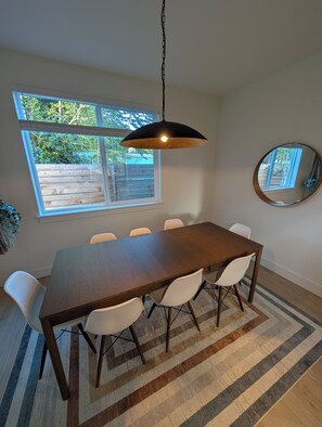 A dining area with a wooden table, white chairs, a hanging pendant light, a large window, and a mirror on the wall. - Live like a local in a brand-new pet-friendly home in the heart of Greenwood. (Seattle)