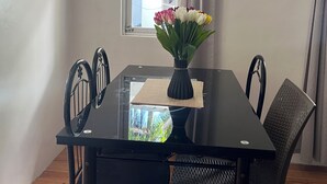 Ruokailutila, jossa on lasipöytä, mustat tuolit, maljakko kukkineen, ikkuna verhoineen ja näkymä vehreään maisemaan.