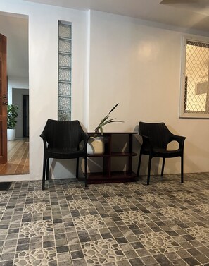 A room with a tiled floor, two wicker chairs, a small table with a plant, a vertical glass panel, and a window with a metal screen.