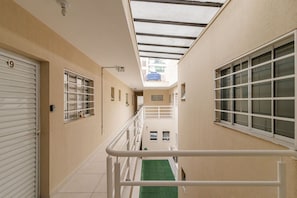 Um corredor estreito com uma clarabóia, uma área externa verde e um corrimão branco.