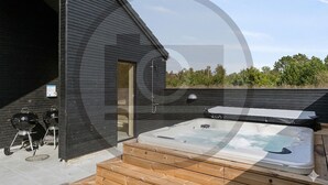 A modern outdoor hot tub area with a wooden deck, a grill, and a door leading to a covered space. - Gorgeous home in Rødby with sauna (Rødby)