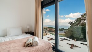 Une chambre d’hôtel avec un lit, une table de chevet, une lampe et une grande fenêtre donnant sur la mer et les bâtiments.