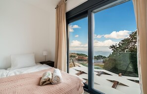 Ein Hotelzimmer mit Bett, Nachttisch, Lampe und einem großen Fenster mit Blick auf das Meer und Gebäude.