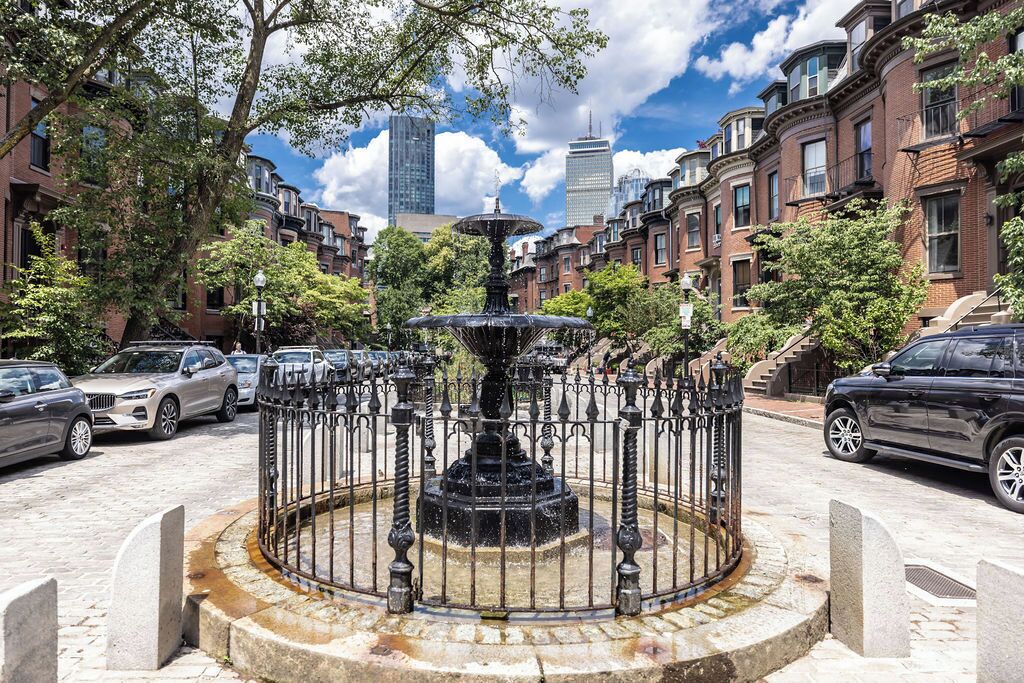 A fountain in a circular stone basin surrounded by a metal fence, with brick buildings and parked cars in the background.