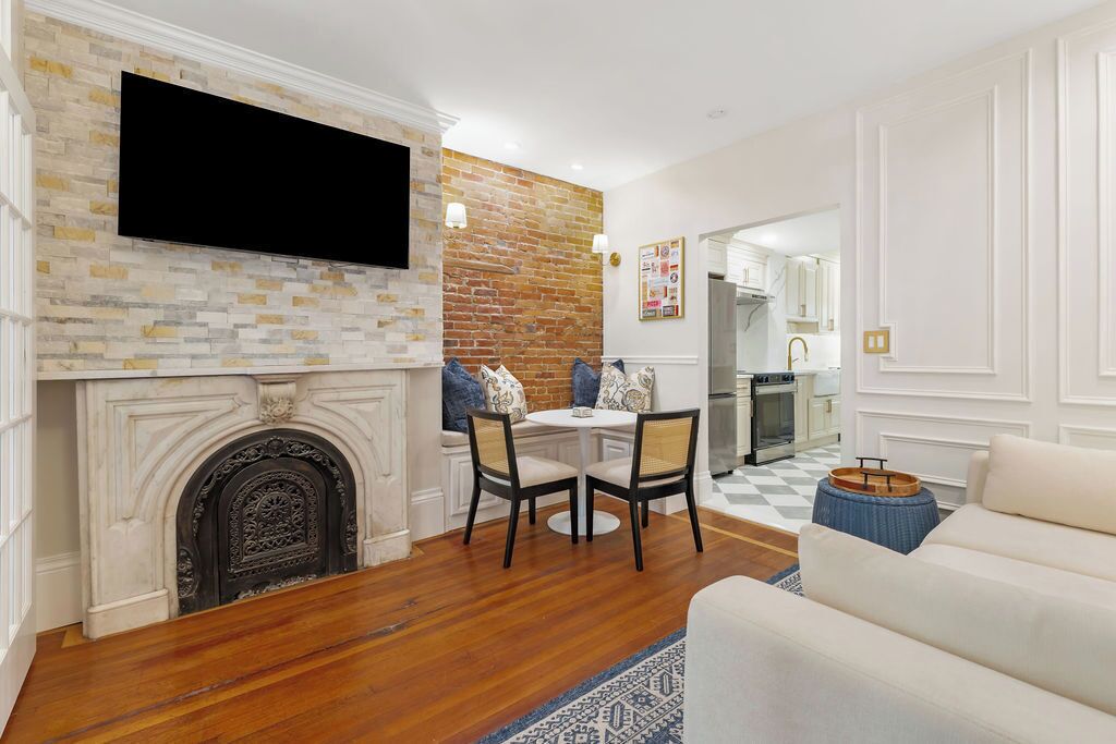 A living room with a fireplace, a television mounted on a brick wall, a dining area with a round table and chairs, and a kitchen in the background.