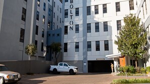 A multi-story building with 'THE BOLTON' signage, a white pickup truck parked in front, and a clear sky. - Luxury Rosebank Apartment  - Pool - Cinema - Backup Power - Secure Parking (Johannesburg)