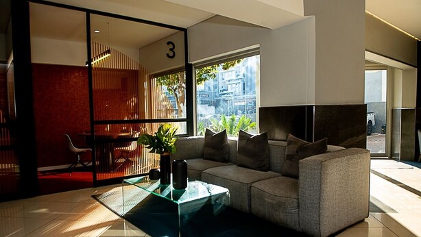A modern living room with a sectional sofa, a glass coffee table, and a view of the city through large windows. - Luxury Rosebank Apartment  - Pool - Cinema - Backup Power - Secure Parking (Johannesburg)