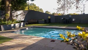 A swimming pool with a tiled deck, surrounded by a low wall and greenery. - Luxury Rosebank Apartment  - Pool - Cinema - Backup Power - Secure Parking (Johannesburg)