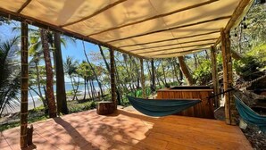 A wooden deck with a canopy, a hammock, and a wooden structure, overlooking a beach with palm trees.