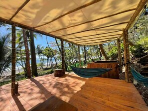 Eine Holzterrasse mit Sonnensegel, einer Hängematte und einer Holzhütte, mit Blick auf einen Strand mit Palmen.