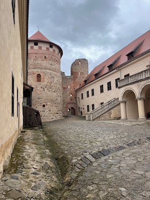 A historic stone courtyard with a castle tower, a wooden gate, and a building with a staircase.
