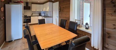 A wooden-paneled room with a dining table, chairs, a kitchen area, and a window with curtains.