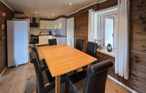 Ein Zimmer mit Holzwänden, einem Esstisch, Stühlen, einer Küchenzeile und einem Fenster mit Vorhängen.