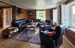 Ein gemütliches Wohnzimmer mit einem schwarzen Ledersofa, einem passenden Sessel, einem Couchtisch und einem Teppich. Ein Fellteppich liegt auf dem Boden, und an der Wand hängt ein Dekorationsobjekt mit einem Bärenkopf.