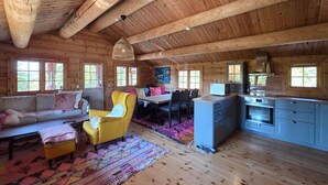 Interior de una cabaña de madera con sofá, sillón amarillo, mesa de comedor, sillas, zona de cocina y ventanas amplias.