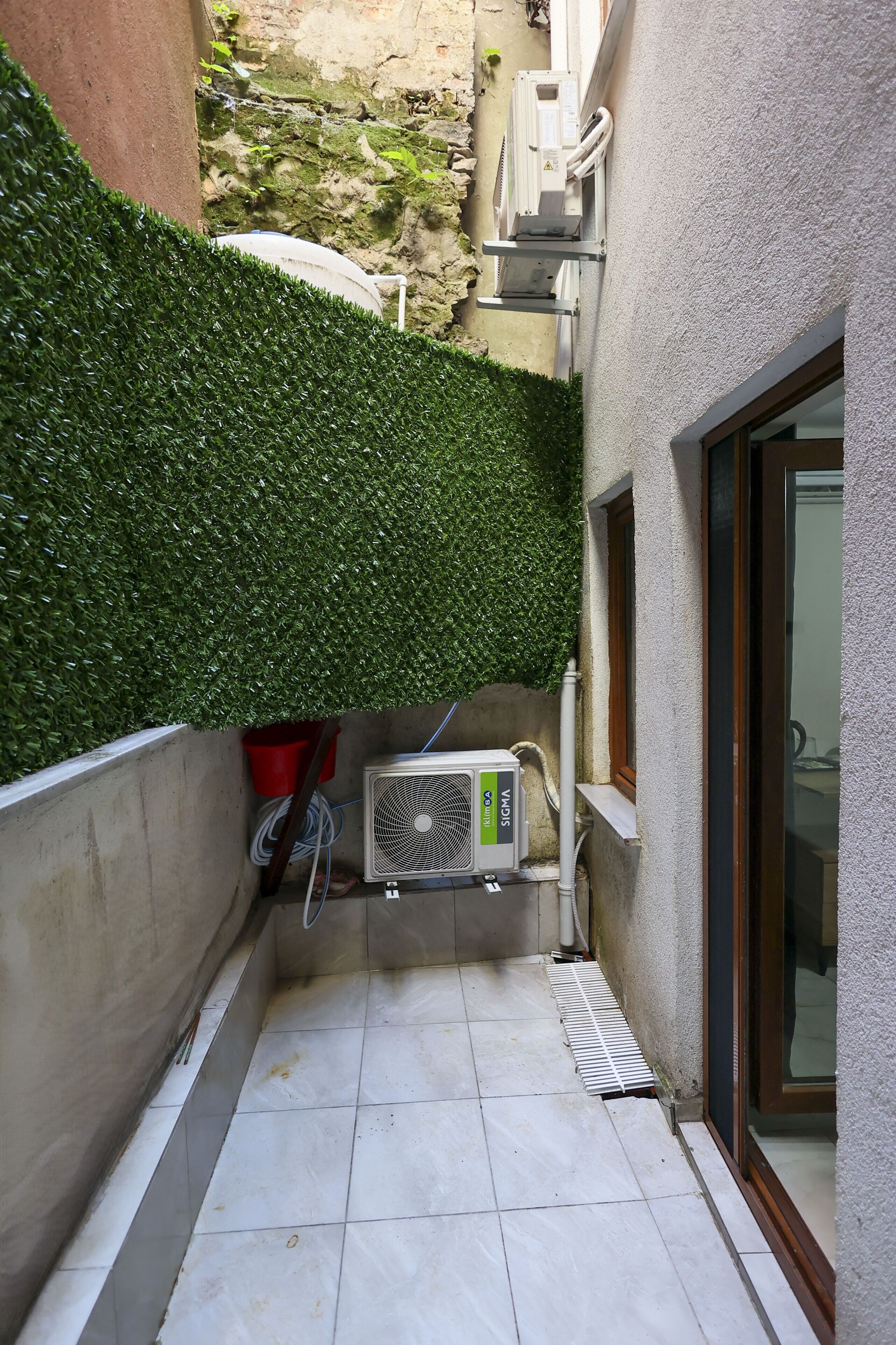 A narrow outdoor corridor with a green hedge, a tiled floor, and an air conditioning unit.