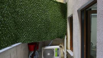 A narrow outdoor corridor with a green hedge, a tiled floor, and an air conditioning unit.