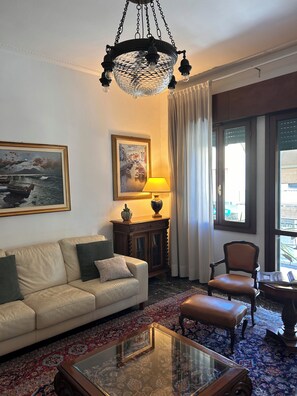 Una sala de estar con sofá, sillón, mesa de centro, candelabro, pinturas, lámpara, aparador y ventanas amplias.