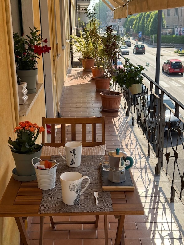 Un balcón con mesa de madera, plantas en macetas, una cafetera y vistas a la calle.