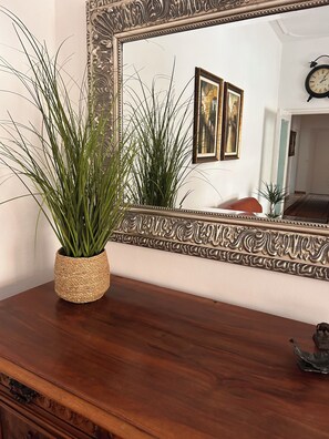 Un comò in legno con una pianta in vaso di vimini, uno specchio con cornice decorata e un orologio appeso al muro.