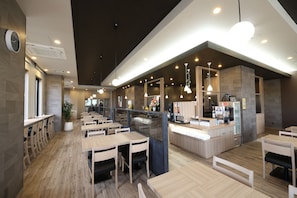 Interior moderno de restaurante com mesas e cadeiras de madeira, balcão com máquina de café e iluminação pendente.
