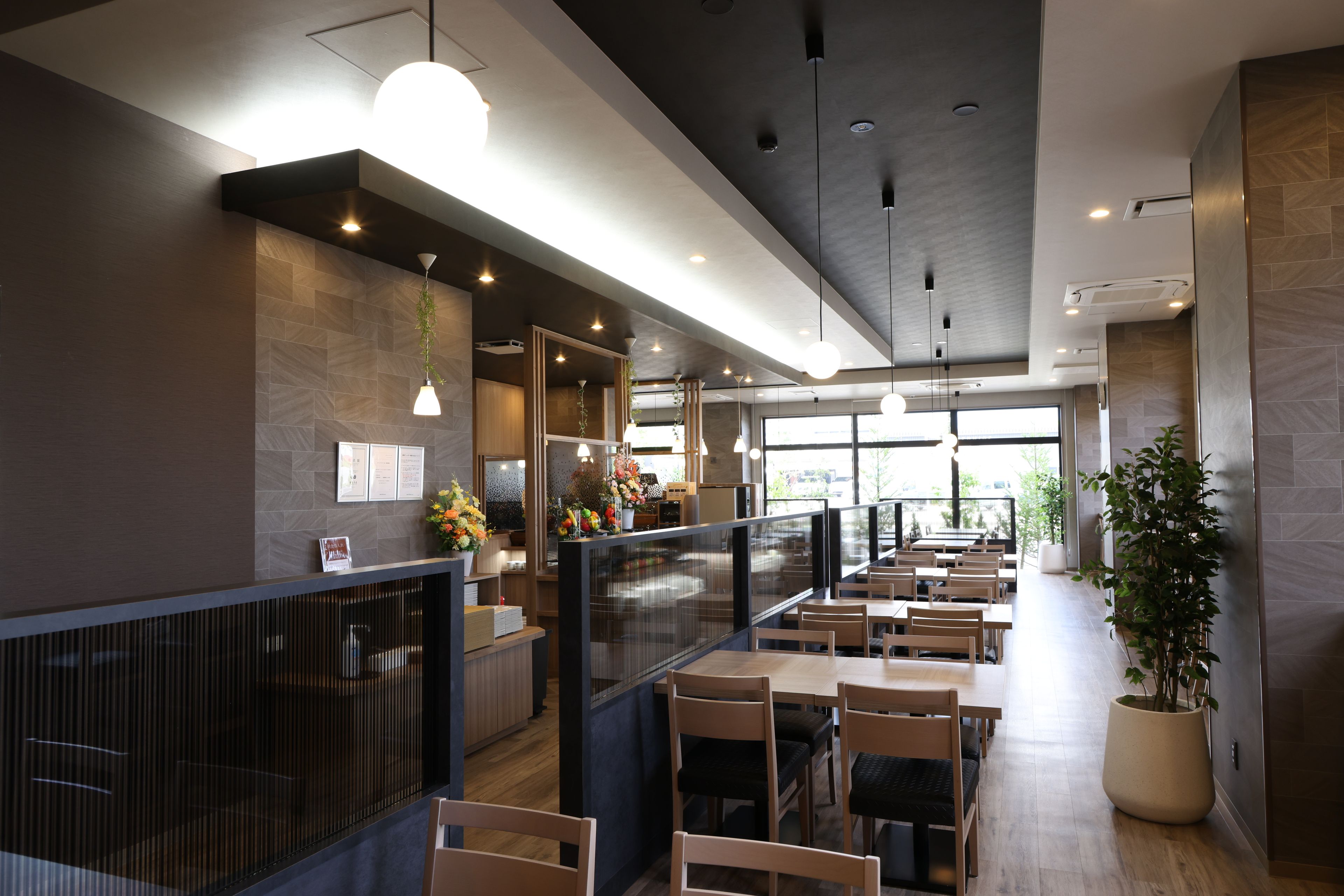 A modern restaurant interior with wooden tables and chairs, hanging pendant lights, and a large potted plant.