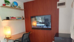 Une pièce avec un téléviseur fixé à un mur rouge, un bureau avec une chaise, une lampe, une petite étagère avec des livres et un globe terrestre, et un réfrigérateur.