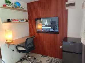 A room with a TV mounted on a red wall, a desk with a chair, a lamp, a small shelf with books and a globe, and a refrigerator.