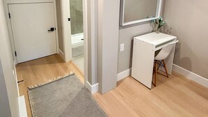 Quarto com piso de madeira, uma escrivaninha branca com cadeira, um espelho e uma porta que dá para outro cômodo.