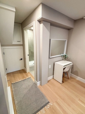 Une pièce avec un plancher en bois, un bureau blanc avec une chaise, un miroir et une porte donnant sur une autre pièce.