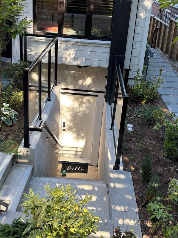 Une entrée d’immeune d’habitation avec un escalier en béton, des rampes en métal, un petit espace jardin et un paillasson avec l’inscription « Bonjour ».
