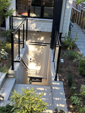 A residential entrance with a concrete staircase, metal railings, a small garden area, and a 'hello' mat.