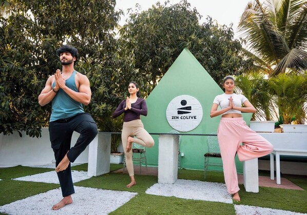 Three people practicing yoga outdoors with a green sign labeled 'ZEN COLIVE' in the background. - Zen Colive (Pune)
