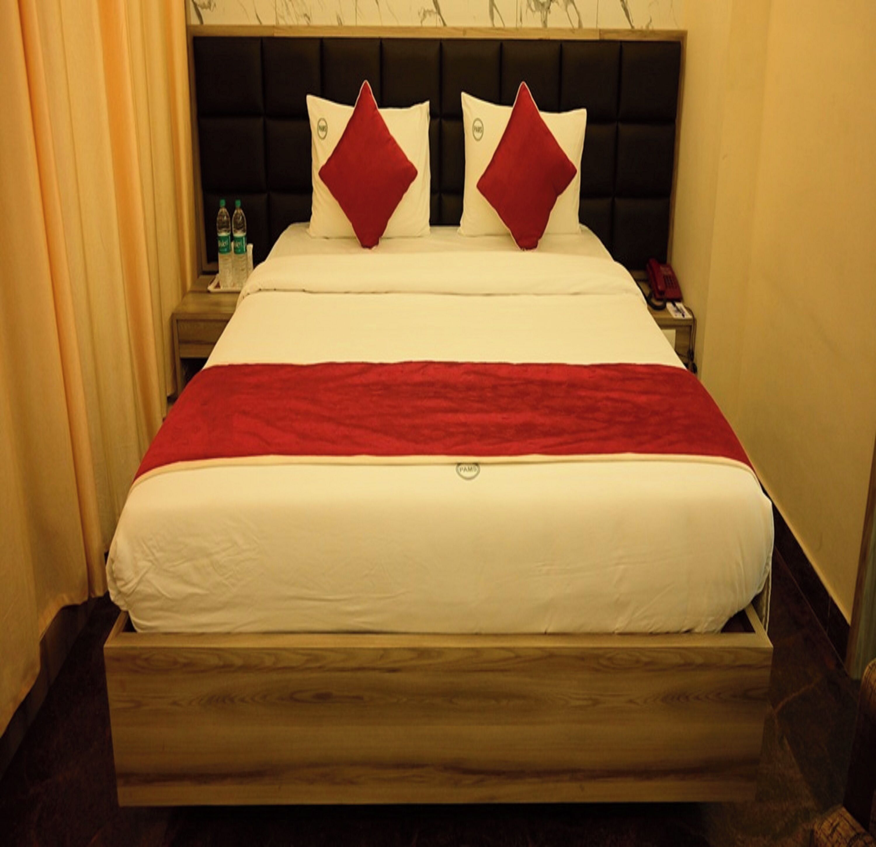 Quarto de hotel com uma cama, duas almofadas vermelhas, cabeceira de madeira, mesa de cabeceira com um telefone e uma garrafa de água.