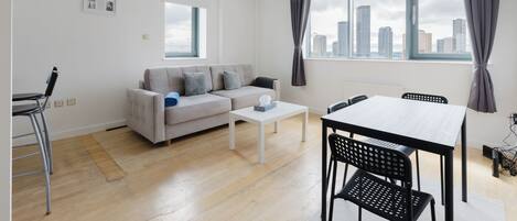 A modern living room with a sofa, coffee table, dining table, and chairs. There are windows with curtains, a radiator, and a city view.