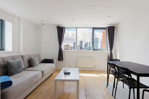 A modern living room with a sofa, a coffee table, a dining table with chairs, and a large window with curtains. - 2BDR Flat with Skyline Views in East London (Greater London)