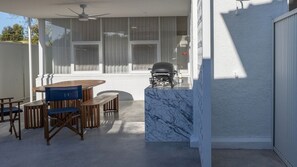 Une terrasse avec une table et des chaises en bois, un barbecue et un mur blanc agrémenté d’un élément décoratif en marbre.