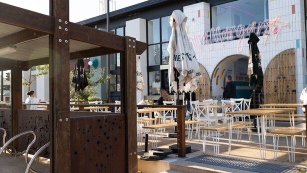 Un café al aire libre con mesas y sillas de madera, una cerca de metal y un edificio con un letrero de neón al fondo.