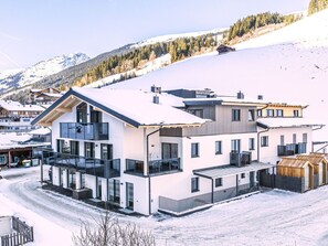A modern multi-story building with large windows, a covered outdoor area, and a view of a snowy mountain landscape. - Spacious apartment with ski in ski out and sauna (Hinterglemm)