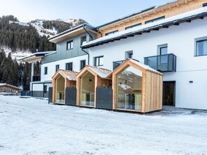 Un edificio moderno con un paisaje de estación de esquí al fondo, que presenta una amplia fachada blanca, varios ventanales y una estructura de madera con techo a dos aguas.