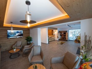 Ein modernes Wohnzimmer mit Fernseher, bequemen Sitzmöglichkeiten, Holzboden und Blick ins Freie.