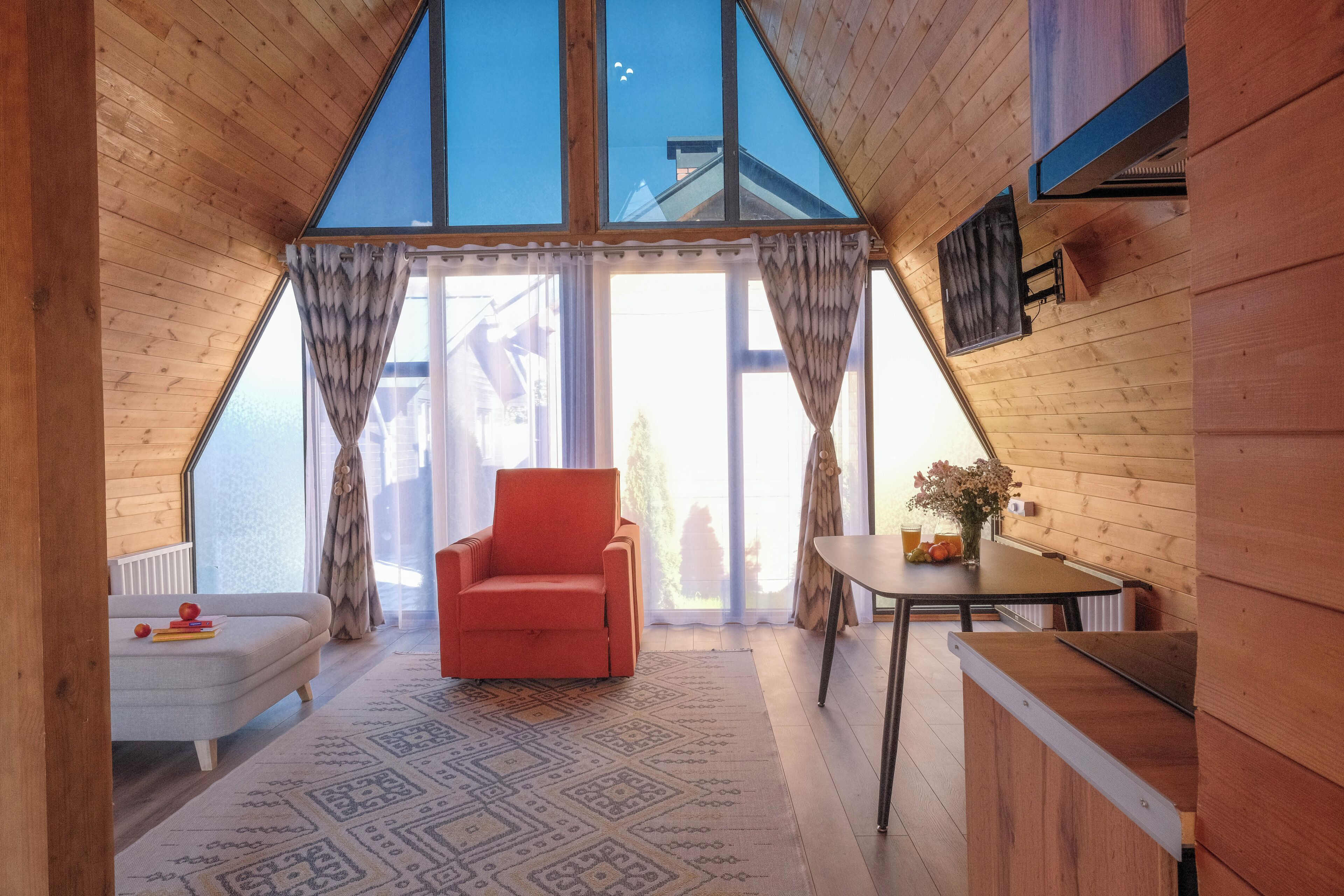 A cozy room with a wooden ceiling, a red chair, a small table with a vase, and a patterned rug.