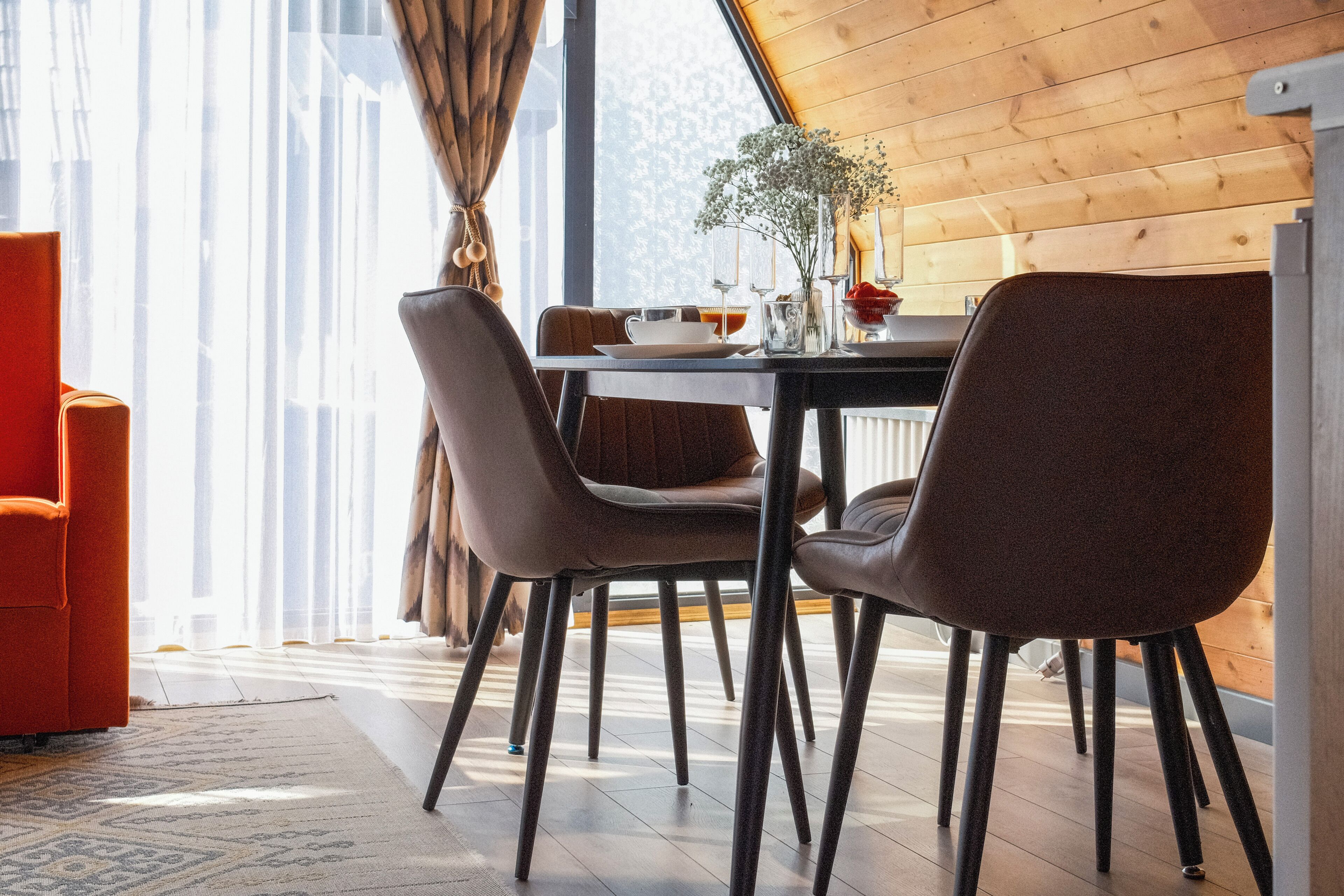 Un comedor con una mesa dispuesta para dos, sillas, un florero con flores y una ventana con cortinas.