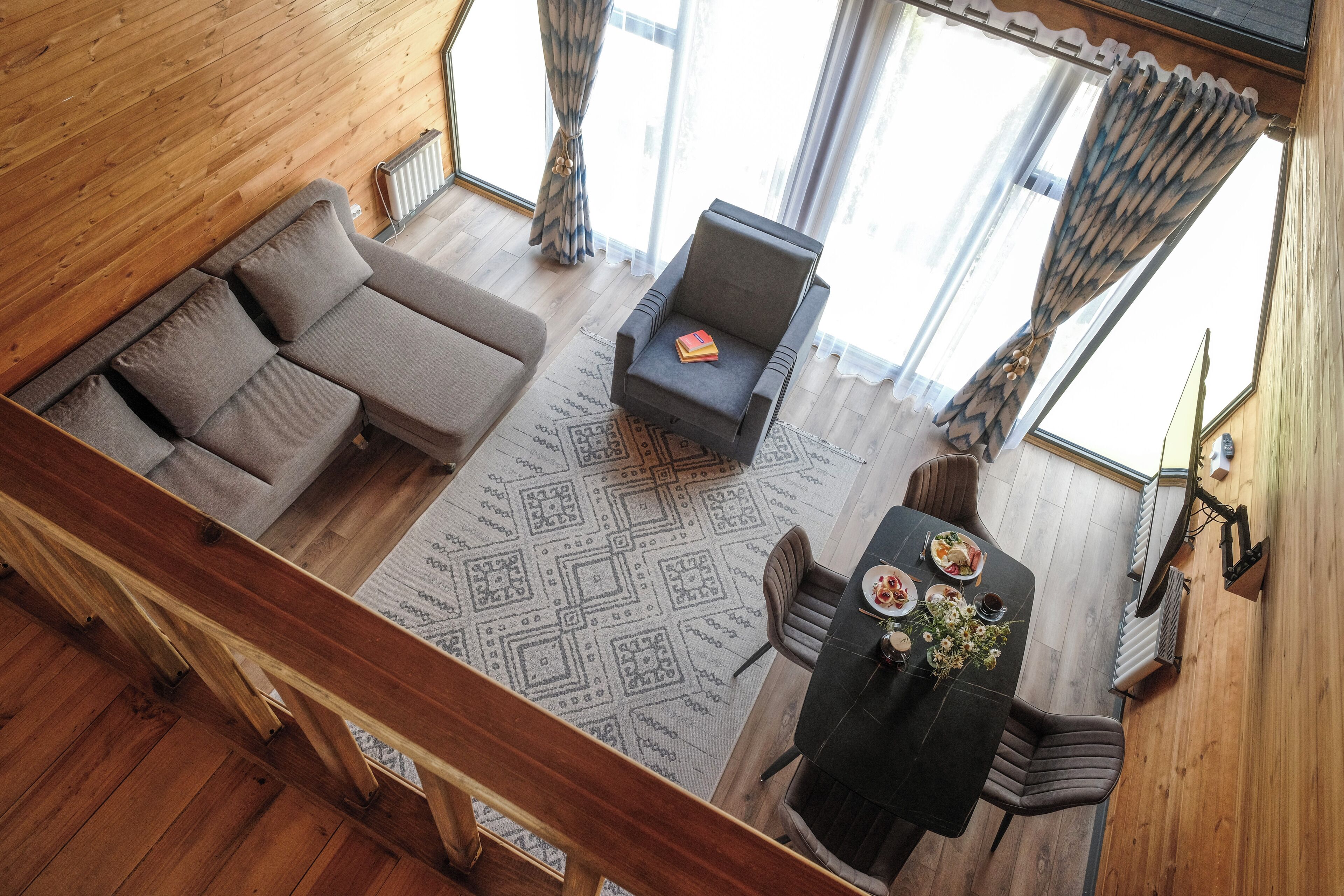 A high-ceiling room with a wooden floor, a sofa, a chair, a dining table with chairs, and a patterned rug.