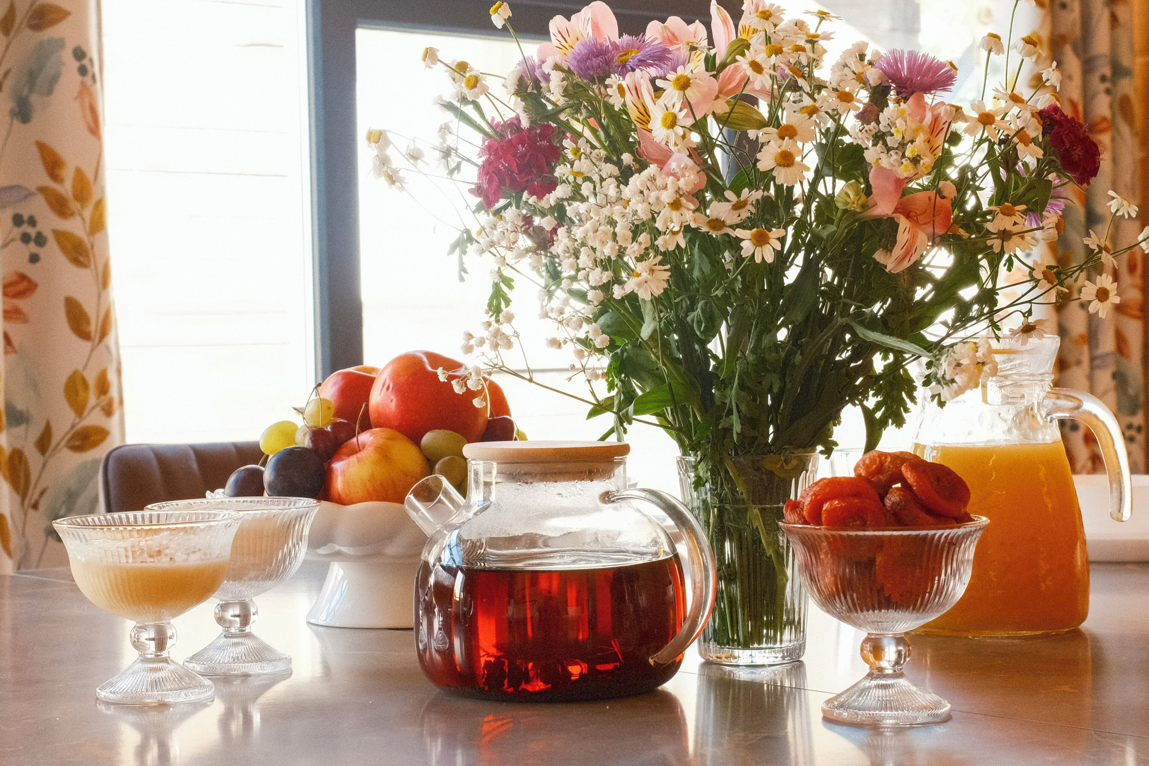 Una mesa con una jarra de té, un vaso de jarra, un cuenco de fruta y un jarrón de flores.