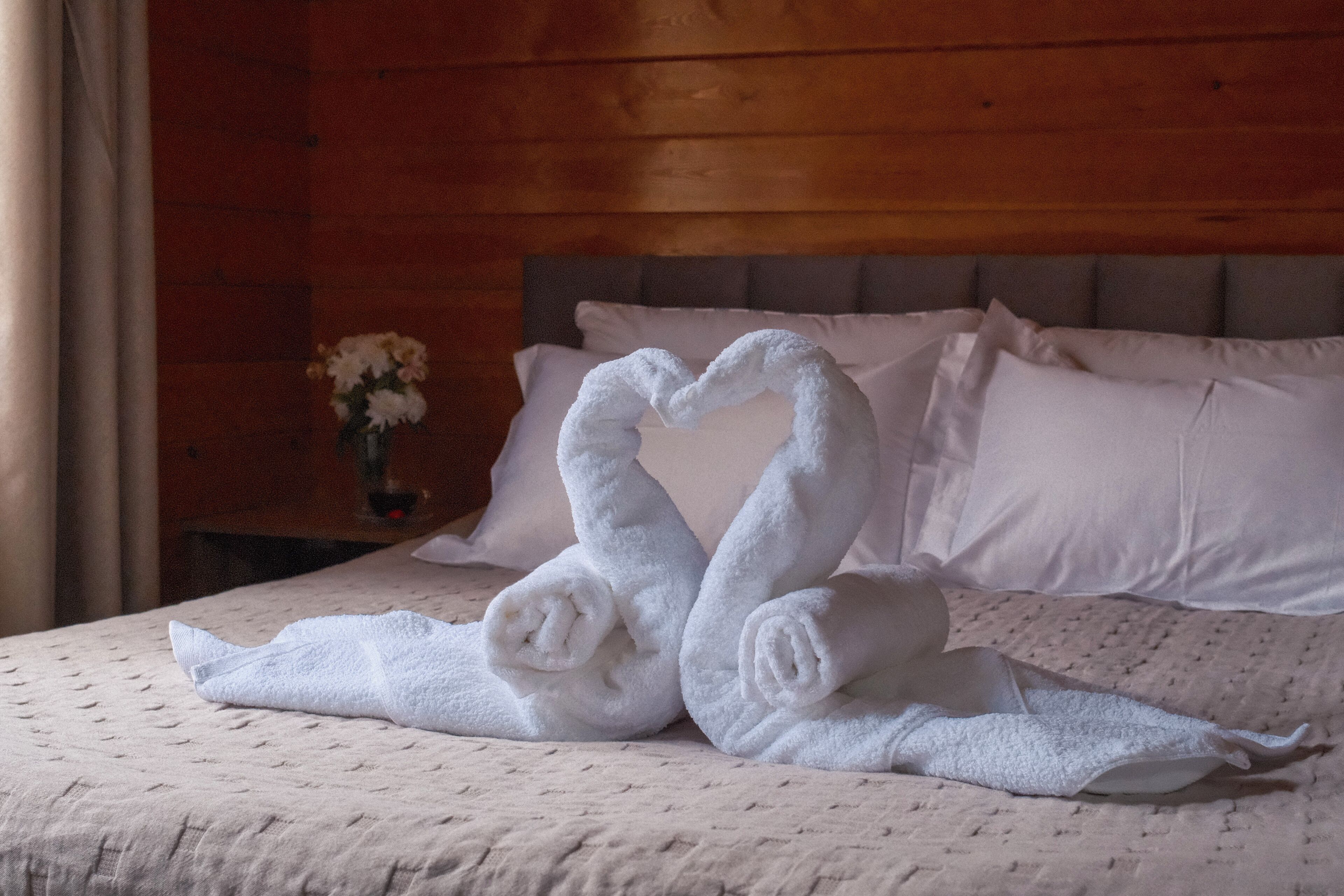 A bed with a heart-shaped towel arrangement, a vase with flowers, and a wooden headboard.